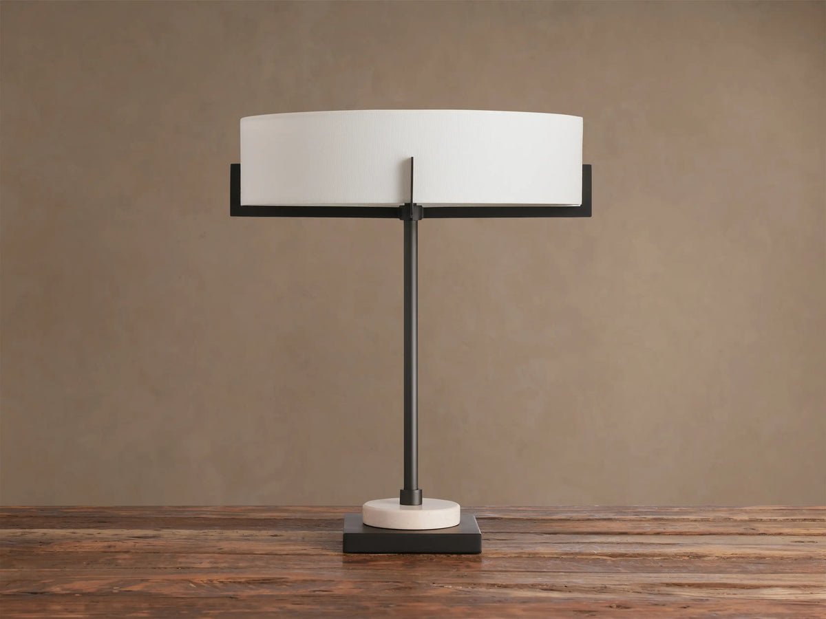 Illuminated Frederic Table Lamp with a bronze frame, white linen shade, and a cream marble disc on a dark square base, on wood.