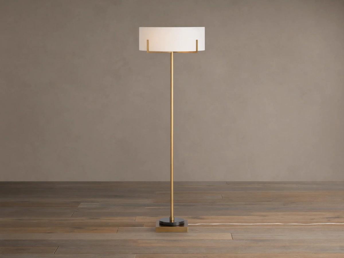 Frederic Floor Lamp: tall antiqued brass pole, white linen shade, black granite base on a wooden floor against a brown wall.
