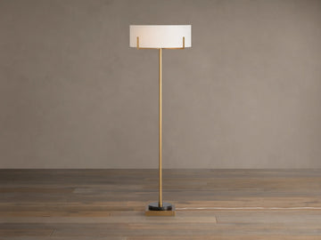 Frederic Floor Lamp: tall antiqued brass pole, white linen shade, black granite base on a wooden floor against a brown wall.