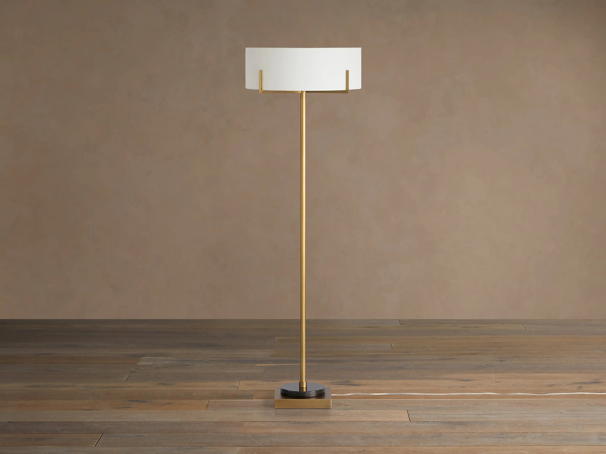 Frederic Floor Lamp: tall antiqued brass pole, white linen shade, black granite base on a wooden floor against a brown wall.