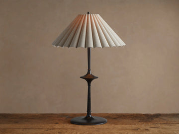 Santiago Table Lamp with a dark bronze base & stem, and a natural pleated linen shade emitting a warm glow, on a wooden table.