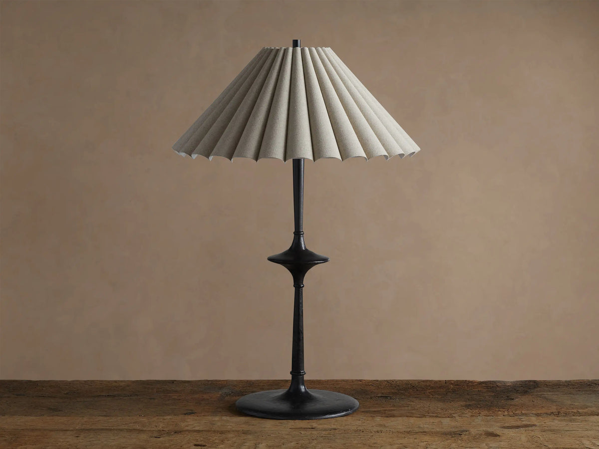 Santiago Table Lamp with a dark bronze base & stem, and a natural pleated linen shade emitting a warm glow, on a wooden table.