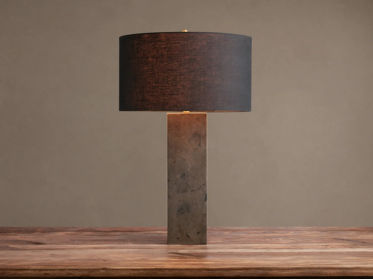 Pyrite Table Lamp with a rectangular base made of dark, shimmering pyrite pieces, topped by a black linen drum shade on a wooden table.