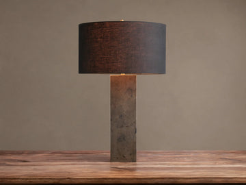 Pyrite Table Lamp with a rectangular base made of dark, shimmering pyrite pieces, topped by a black linen drum shade on a wooden table.