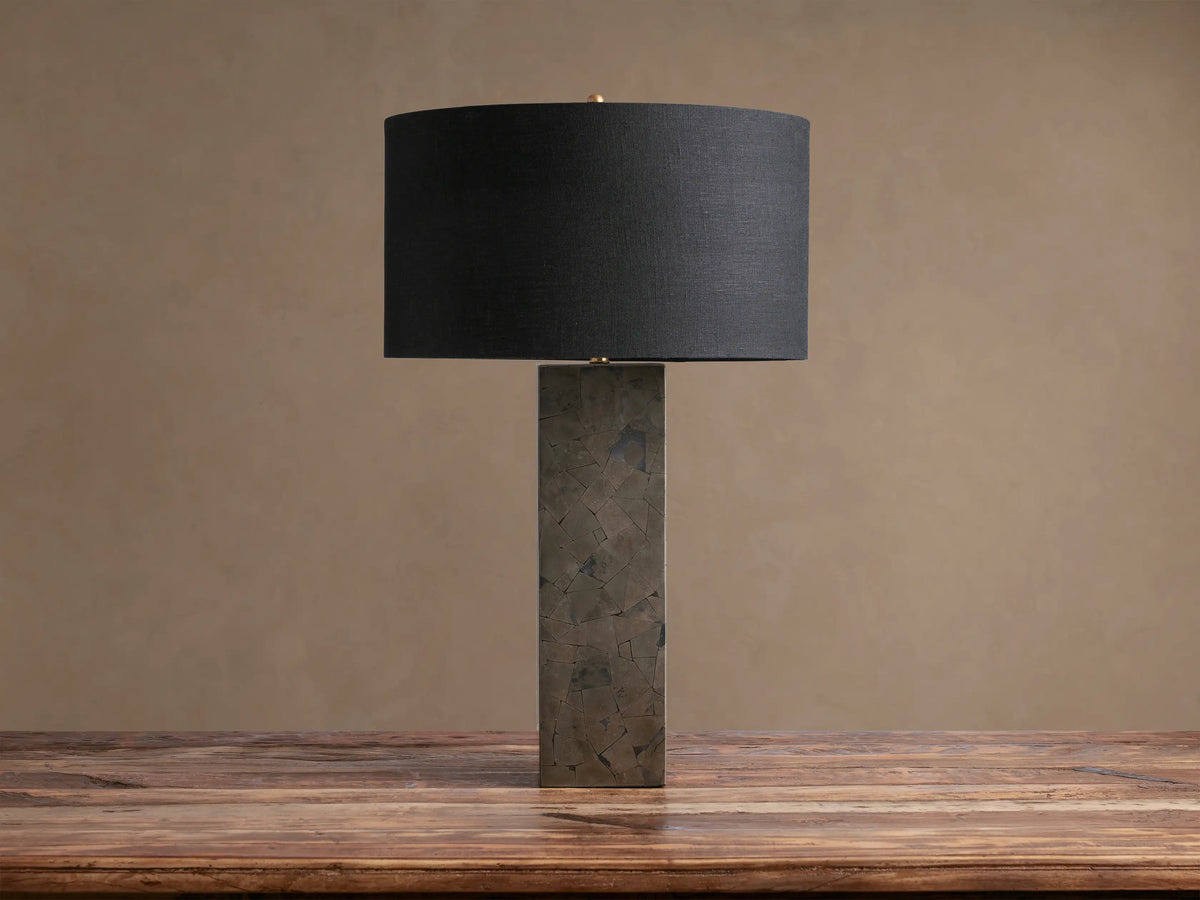 Pyrite Table Lamp with a rectangular base made of dark, shimmering pyrite pieces, topped by a black linen drum shade on a wooden table.