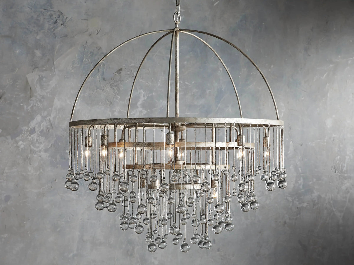 Aubrey Round Chandelier in Silver with a handcrafted iron frame, hanging glass crystals, and five bulbs, set against a grey wall.