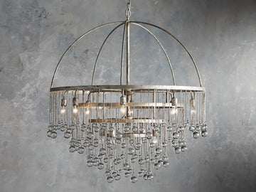 Aubrey Round Chandelier in Silver with a handcrafted iron frame, hanging glass crystals, and five bulbs, set against a grey wall.