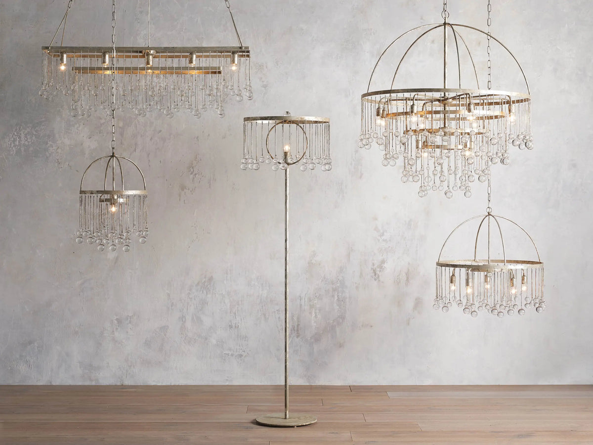 Aubrey Round Chandelier in Silver with a handcrafted iron frame, hanging glass crystals, and five bulbs, set against a grey wall.