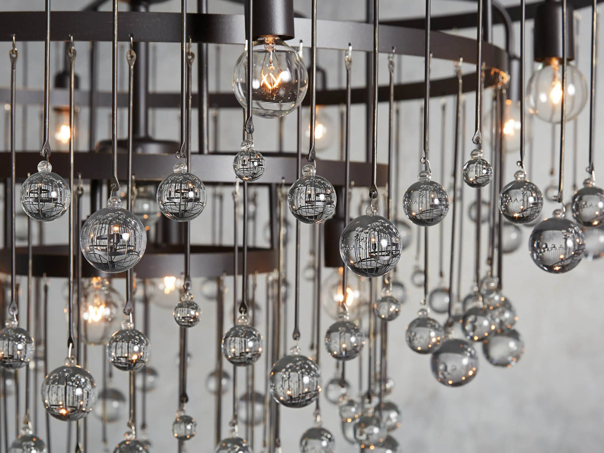 Aubrey Round Chandelier with solid iron frame, multiple tiers, and hanging glass globe accents, set against a textured gray background.