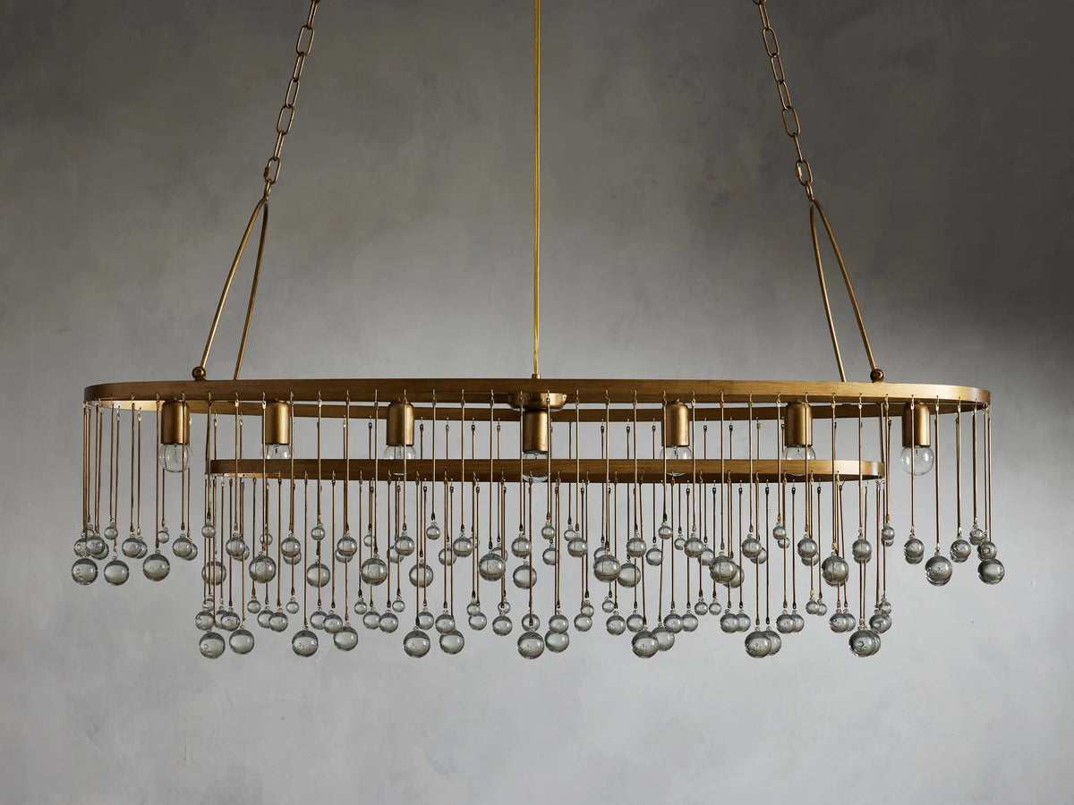 Oval chandelier with gold finish and clear glass droplets, hanging from a double chain. Holds seven bulbs, against a gray background.