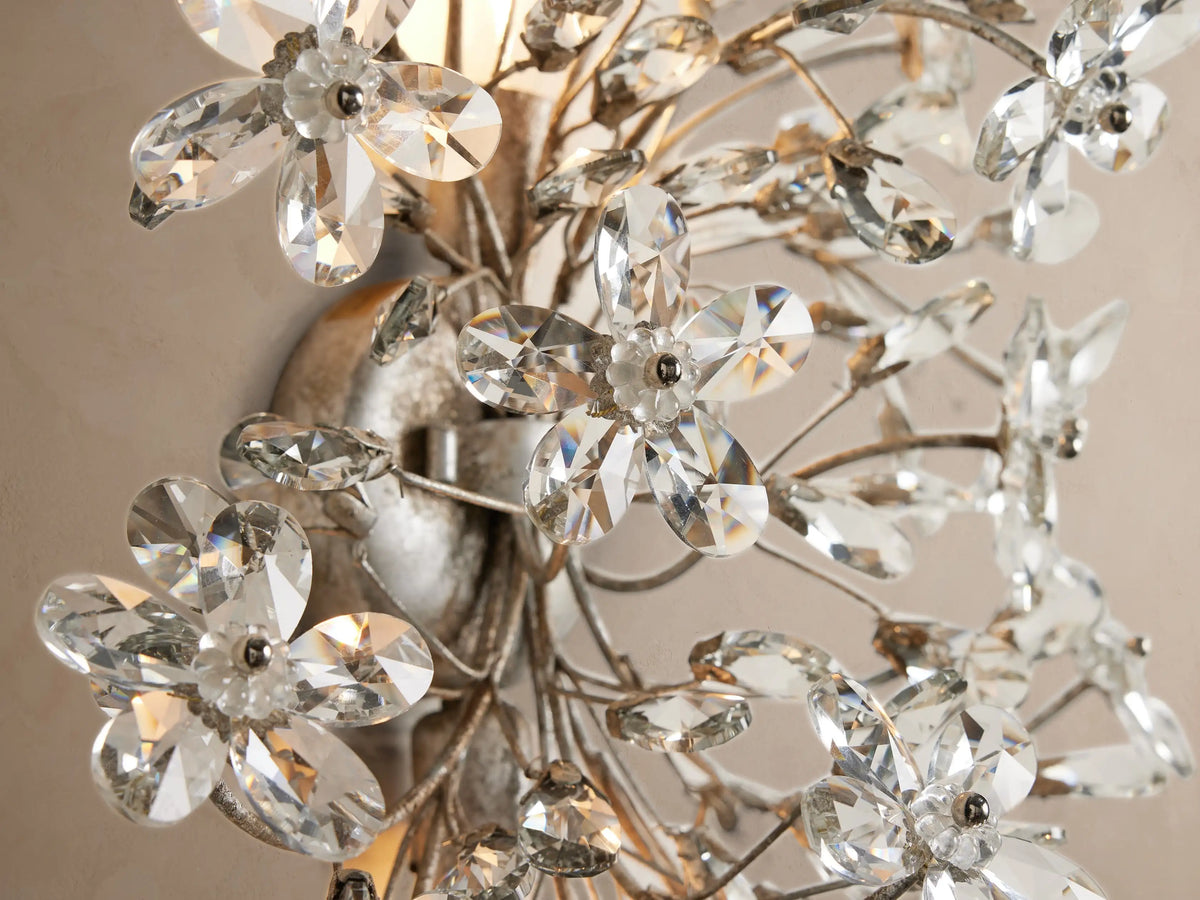 Fiore Sconce with an antiqued silver finish, adorned with numerous sparkling clear crystal flowers and buds, against a beige wall.