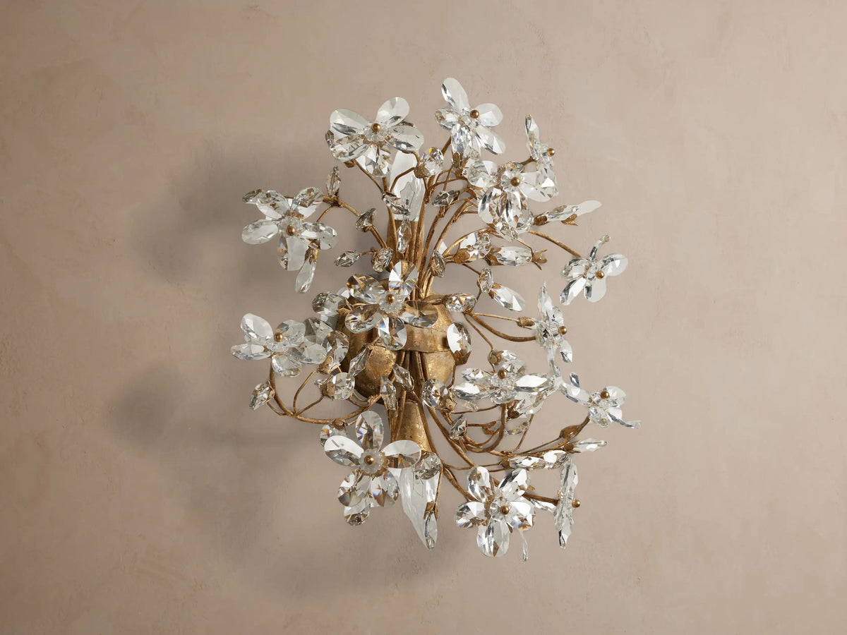 Fiore Sconce: Elegant gold iron sconce with clear crystal flowers, sparkling and glowing with warm light, mounted on a neutral wall.