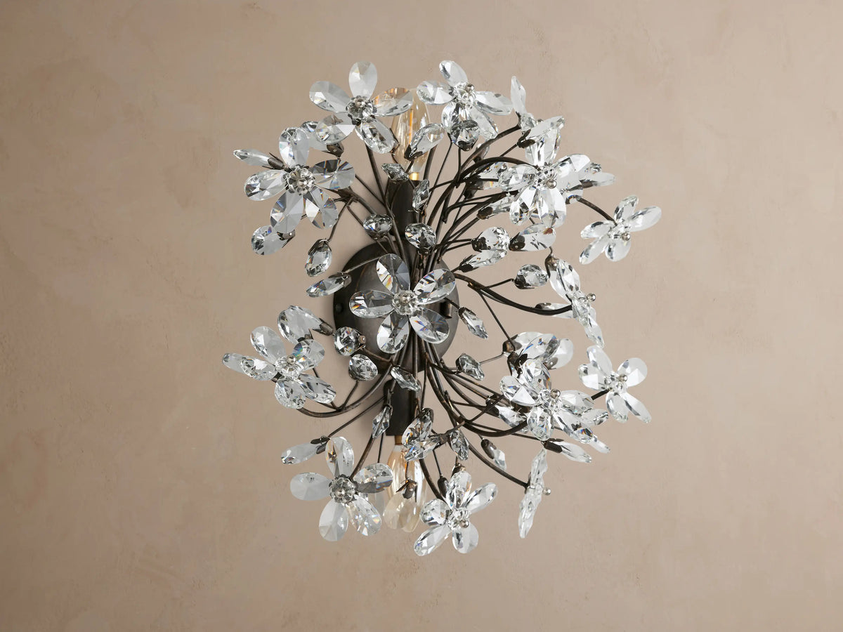 Fiore Sconce, a dark metal wall light with many clear crystal flowers & leaf accents, glowing warmly on a tan wall.