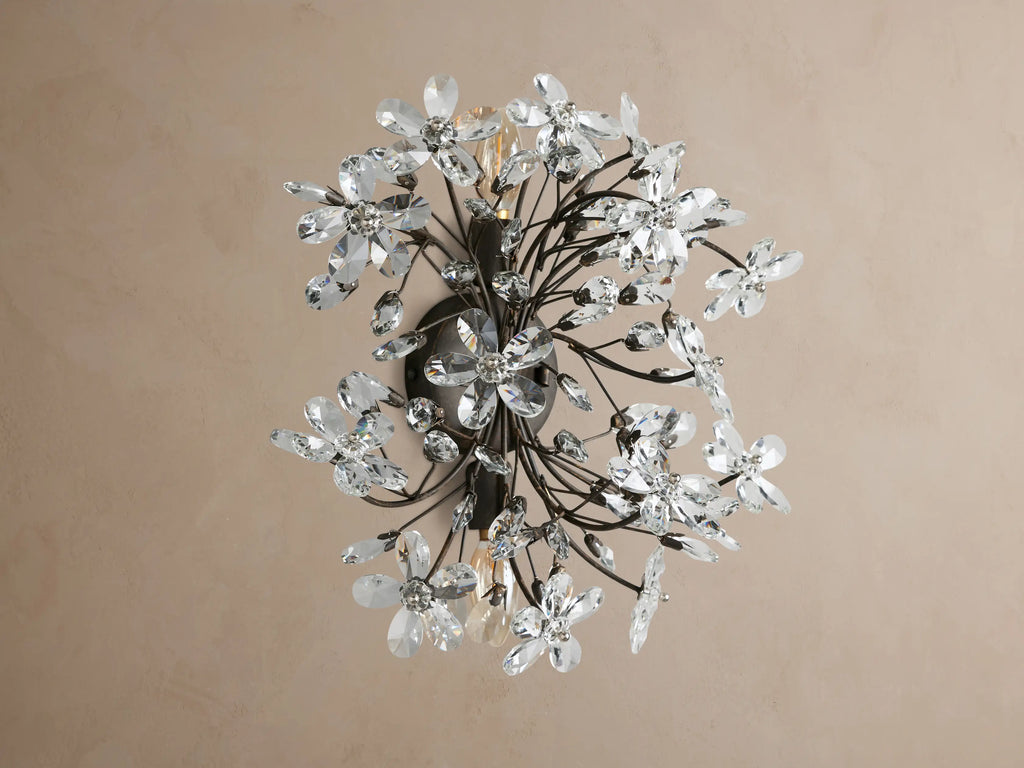 Fiore Sconce, a dark metal wall light with many clear crystal flowers & leaf accents, glowing warmly on a tan wall.
