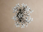 Fiore Sconce, a dark metal wall light with many clear crystal flowers & leaf accents, glowing warmly on a tan wall.