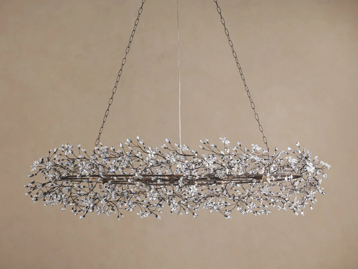 Horizontal Fiore Oval Chandelier, dark iron frame adorned with countless clear crystal floral clusters, glowing warmly.