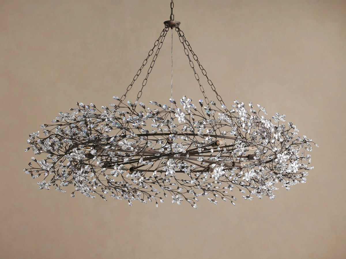 Fiore Crown Chandelier. Oval, dark metal frame covered in sparkling clear crystal flowers, suspended by chains on a light brown background.