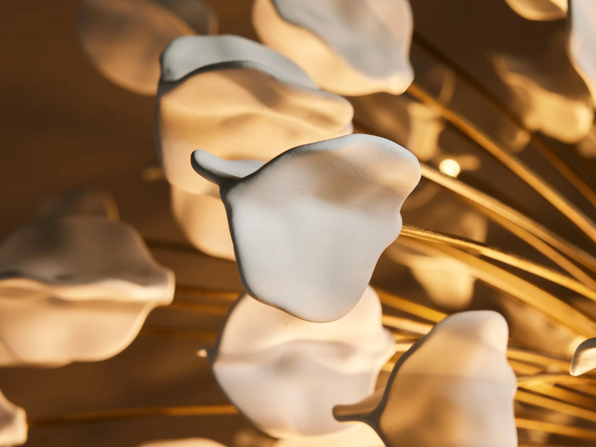 Alt text: Close-up of the Lunaria Chandelier featuring white ceramic leaves on golden iron branches, illuminated by bulbs.