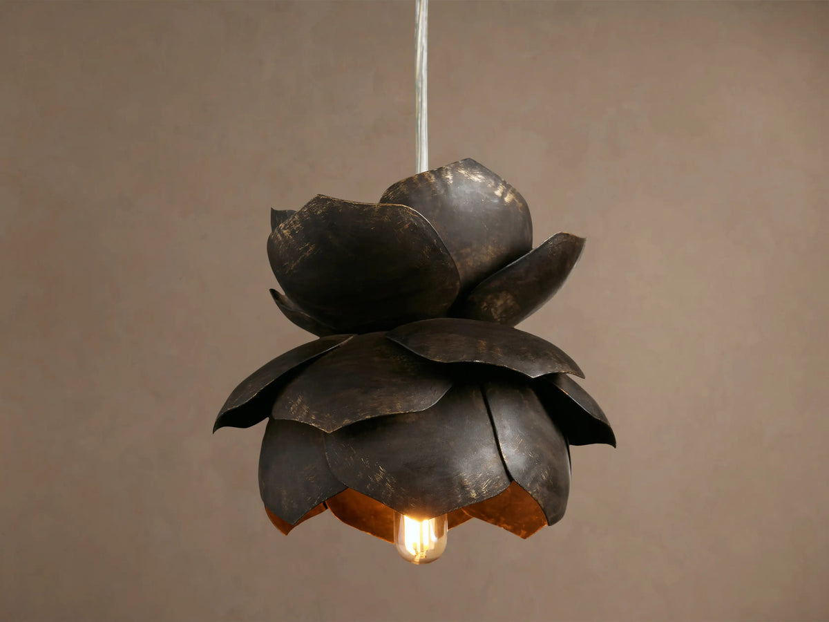 Lorca Pendant, a layered dark bronze metal flower light with distressed brass accents and a warm glowing bulb against a brown wall.