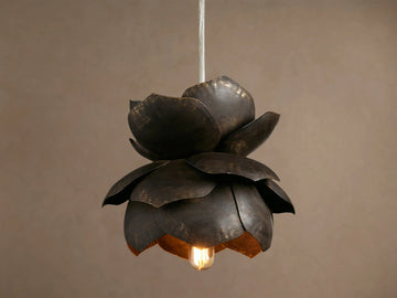 Lorca Pendant, a layered dark bronze metal flower light with distressed brass accents and a warm glowing bulb against a brown wall.