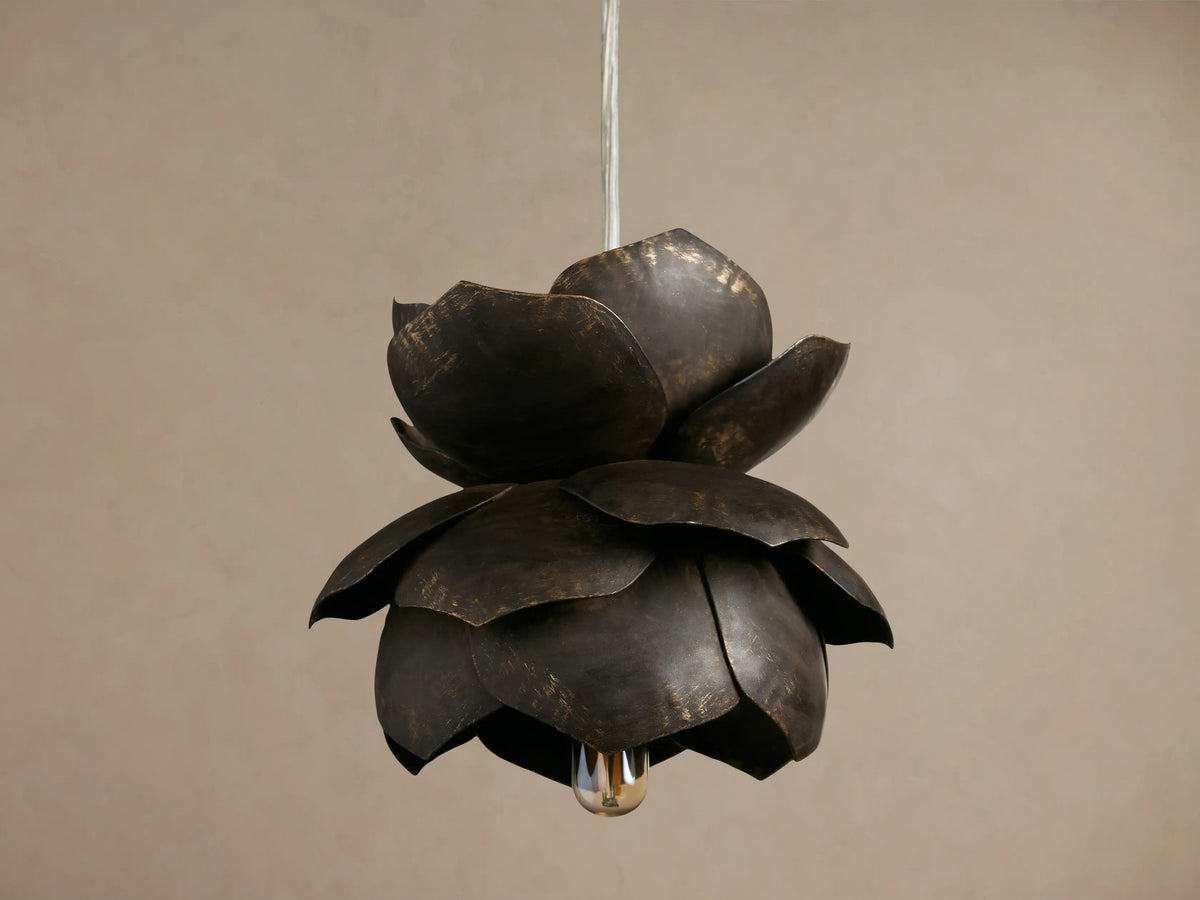 Lorca Pendant, a layered dark bronze metal flower light with distressed brass accents and a warm glowing bulb against a brown wall.