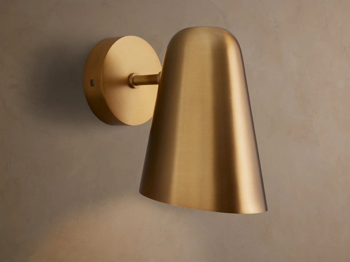 Elih Sconce, a brushed brass wall-mounted light fixture with a circular backplate and a downward-facing conical shade on a textured wall.