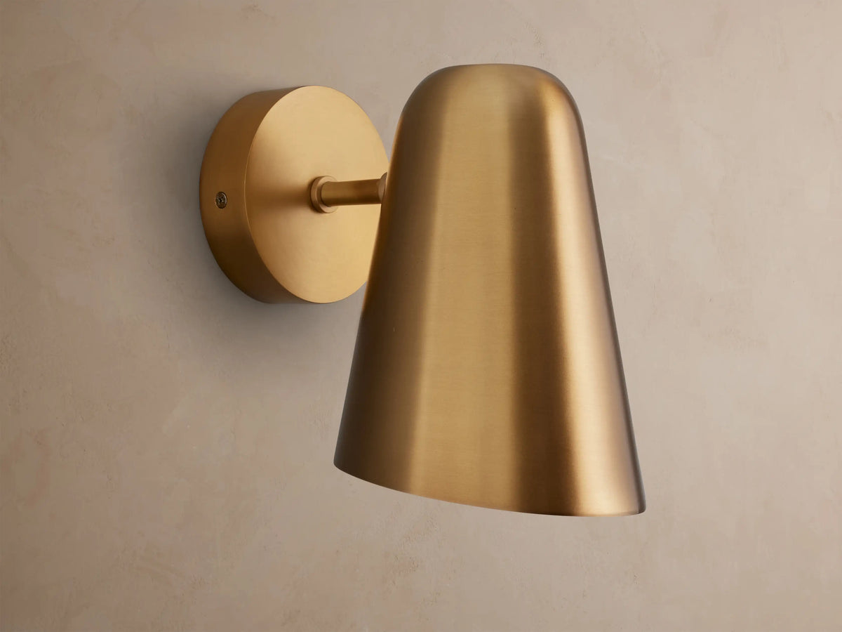 Elih Sconce, a brushed brass wall-mounted light fixture with a circular backplate and a downward-facing conical shade on a textured wall.