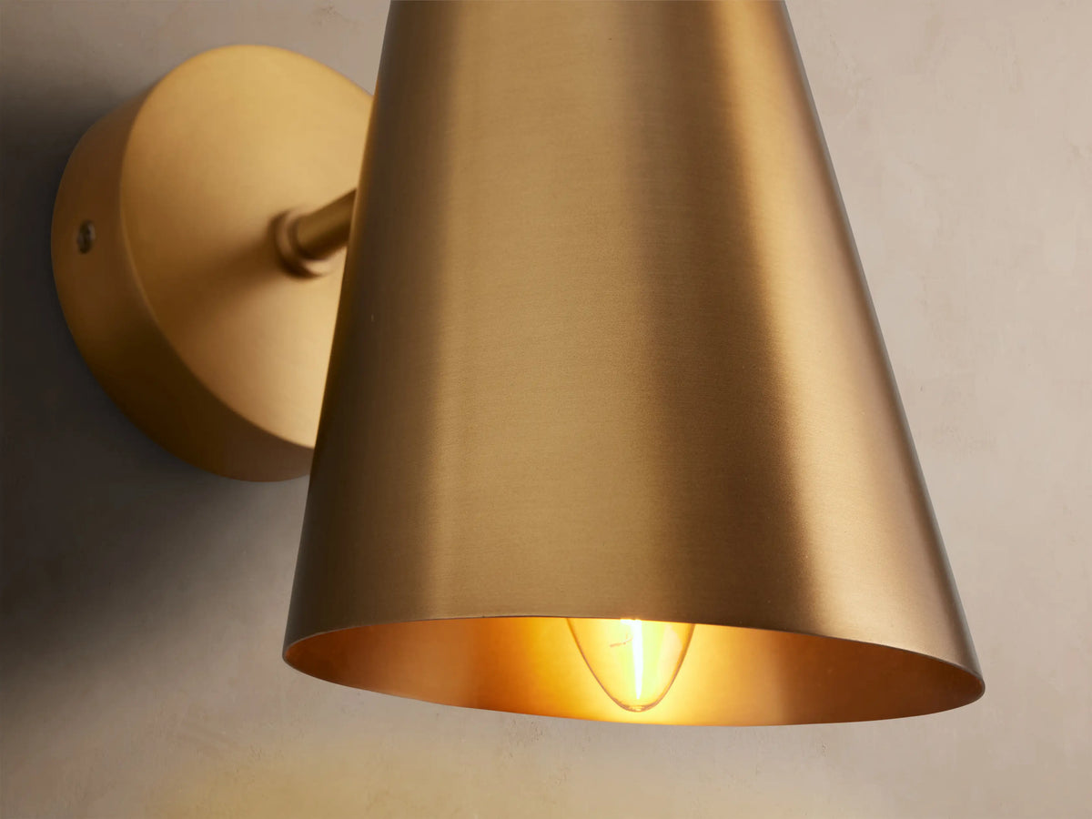 Elih Sconce, a modern wall-mounted light with a brushed brass finish, features a round backplate and a conical shade.