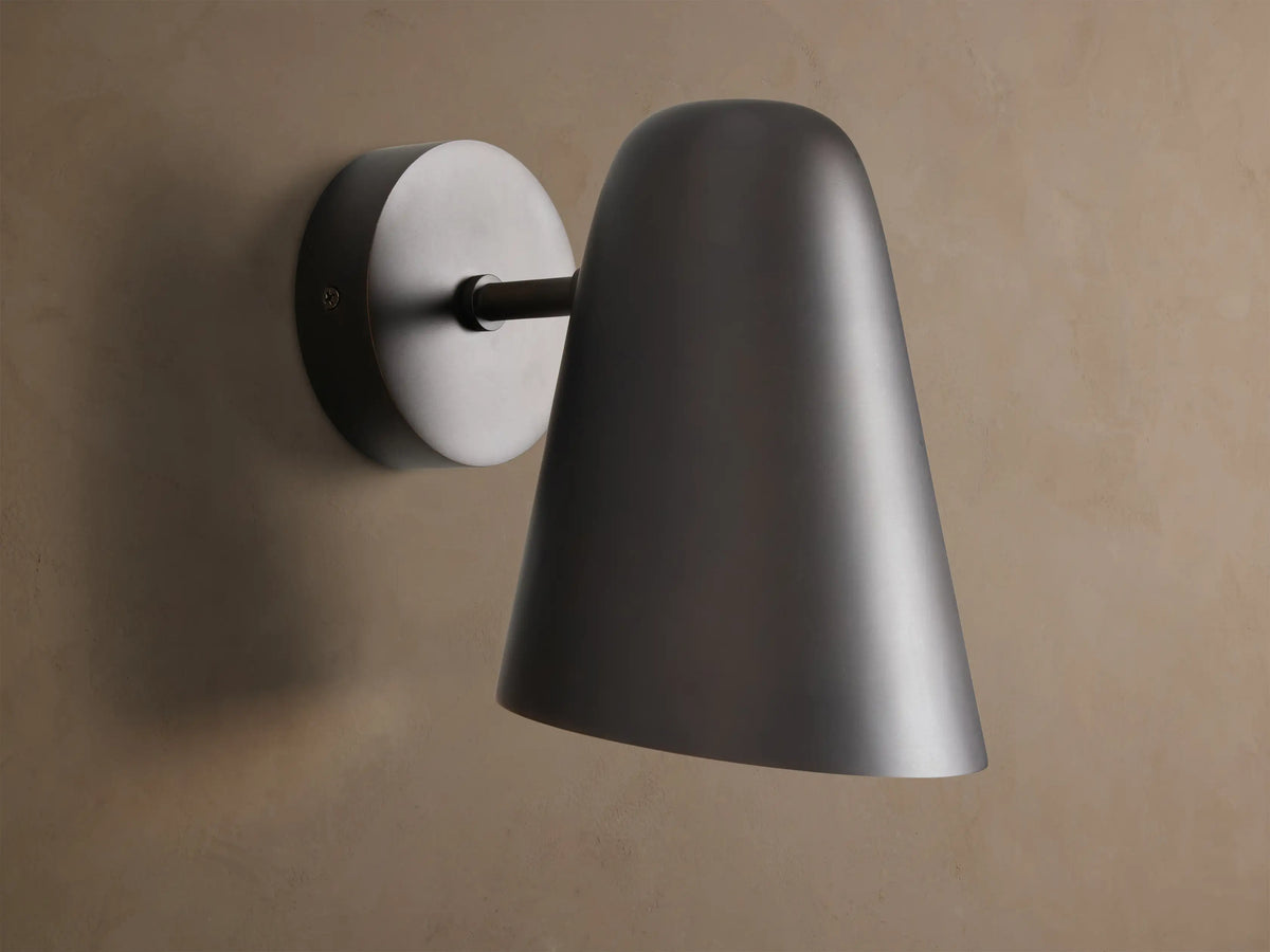 Elih Sconce in antiqued bronze, featuring a conical shade and round backplate, mounted on a textured beige wall.