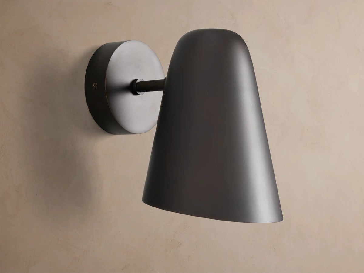 Elih Sconce in antiqued bronze, featuring a conical shade and round backplate, mounted on a textured beige wall.