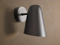 Elih Sconce in antiqued bronze, featuring a conical shade and round backplate, mounted on a textured beige wall.