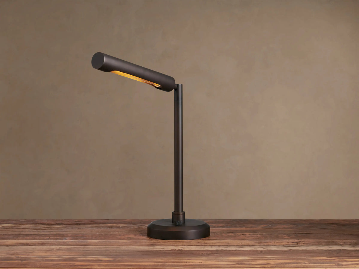 Elih Desk Lamp: Antiqued bronze brass finish, modern design with cylindrical head angled down, emitting warm light on a wooden surface.