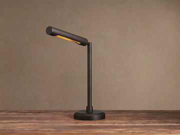 Elih Desk Lamp: Antiqued bronze brass finish, modern design with cylindrical head angled down, emitting warm light on a wooden surface.