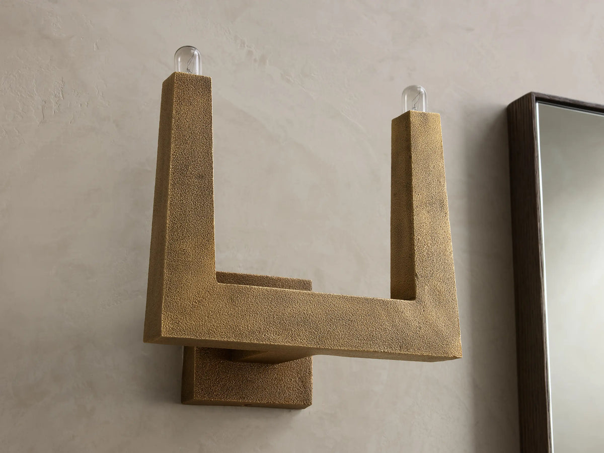 Golden textured wall sconce with an L-shaped arm and a lit bulb at the top, casting warm light on the wall.