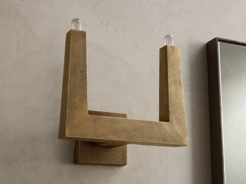 Golden textured wall sconce with an L-shaped arm and a lit bulb at the top, casting warm light on the wall.