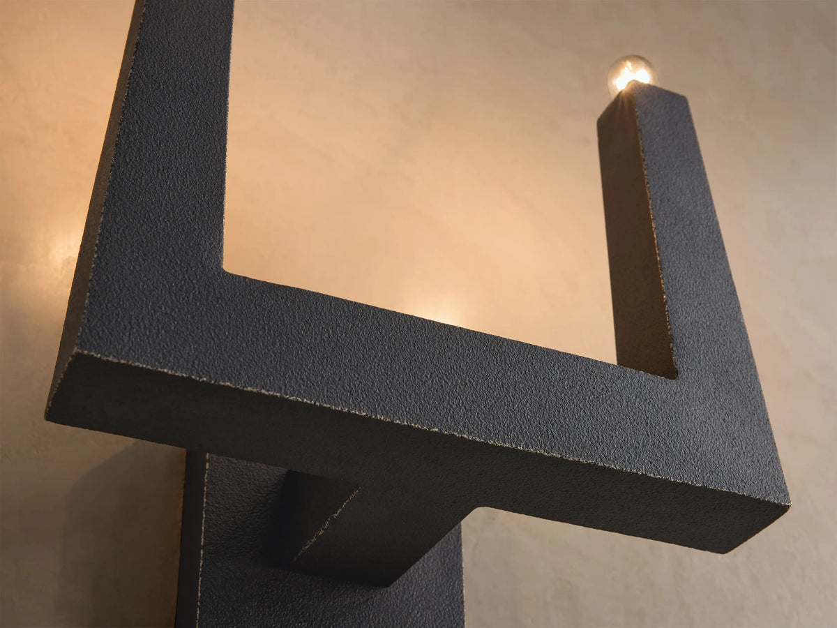 Close-up, angled view of a dark gray textured geometric light fixture with a small glowing bulb at the top right.