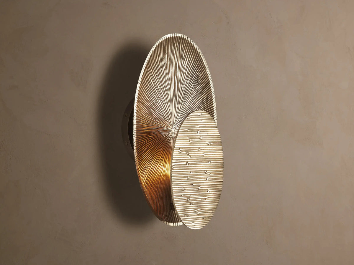 Udaya Embossed Sconce, golden, with overlapping oval plates; one radial, one horizontal embossed. Warm light on a wall.