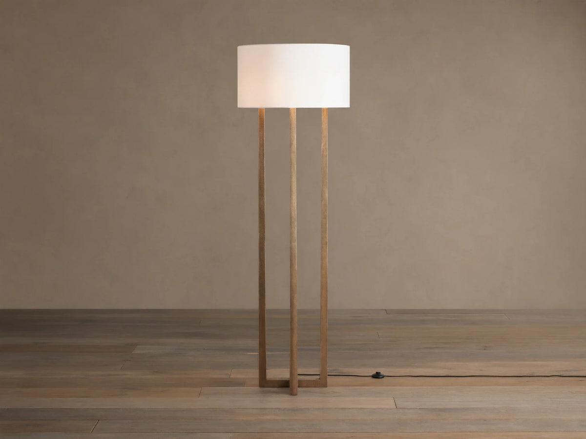 Emmet Floor Lamp with a white linen drum shade & three antiqued brass/bronze vertical columns, standing on a wood floor.