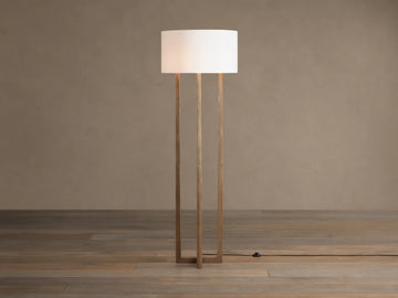 Emmet Floor Lamp with a white linen drum shade & three antiqued brass/bronze vertical columns, standing on a wood floor.