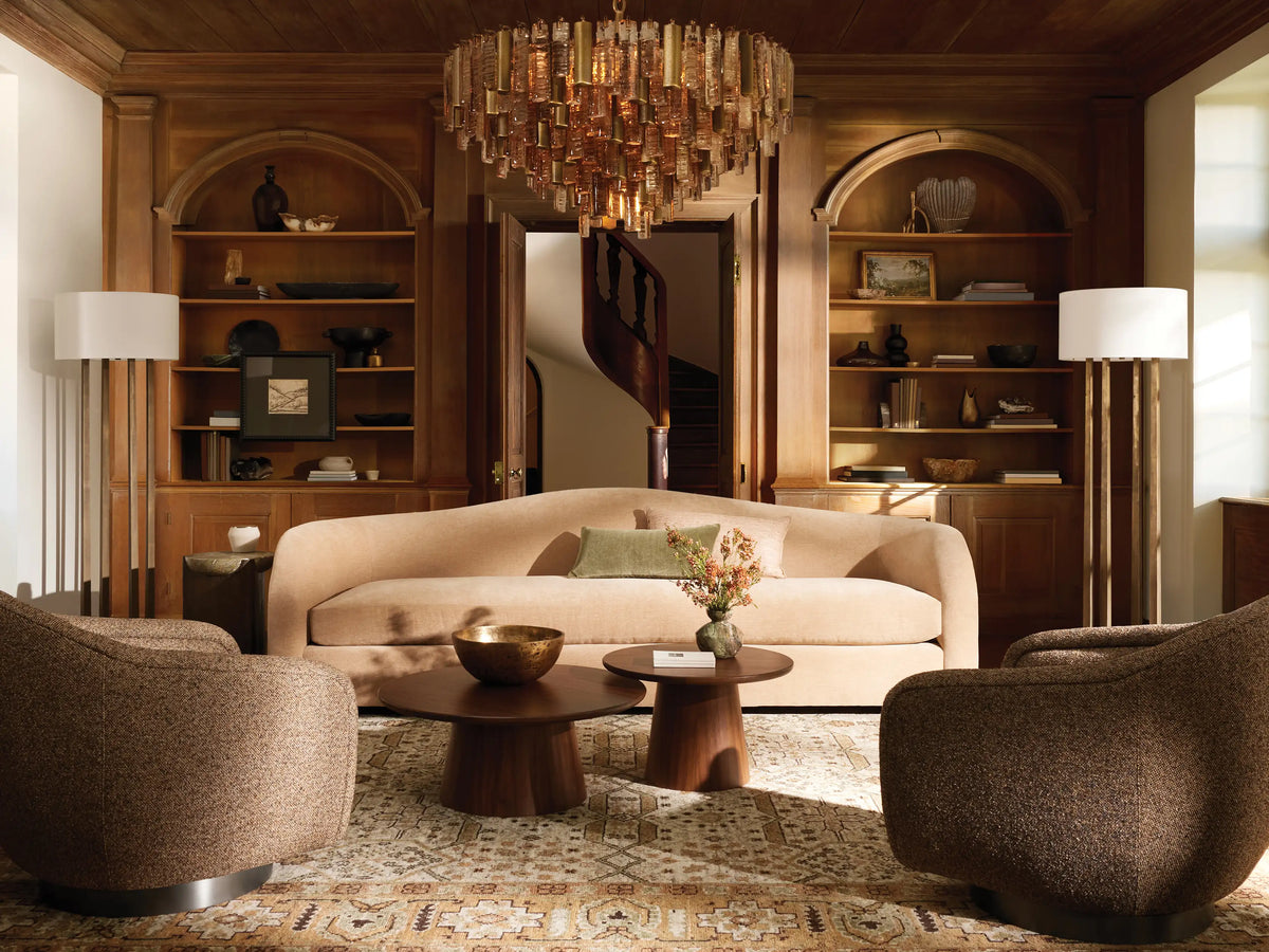 Emmet Floor Lamp with a white linen drum shade & three antiqued brass/bronze vertical columns, standing on a wood floor.