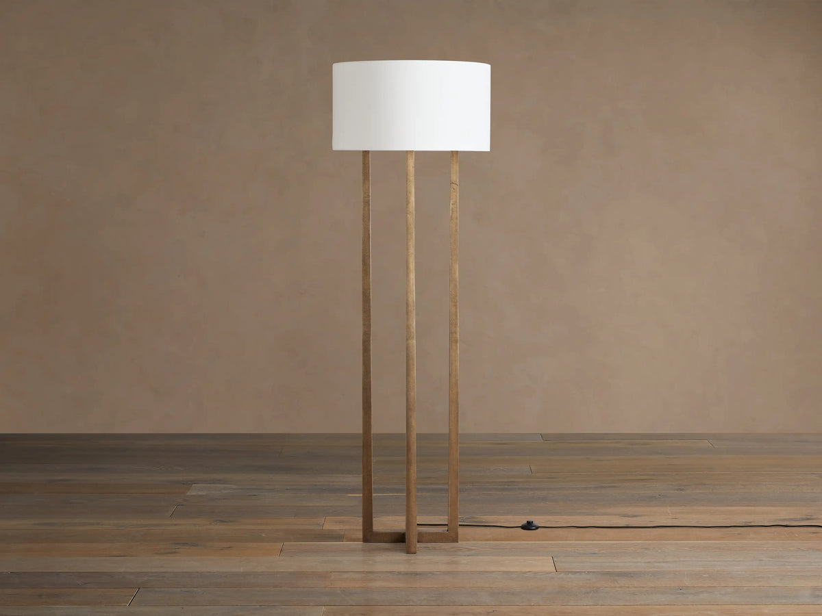 Emmet Floor Lamp with three antiqued brass rectangular legs and white linen drum shade, on wooden floor against a brown wall.