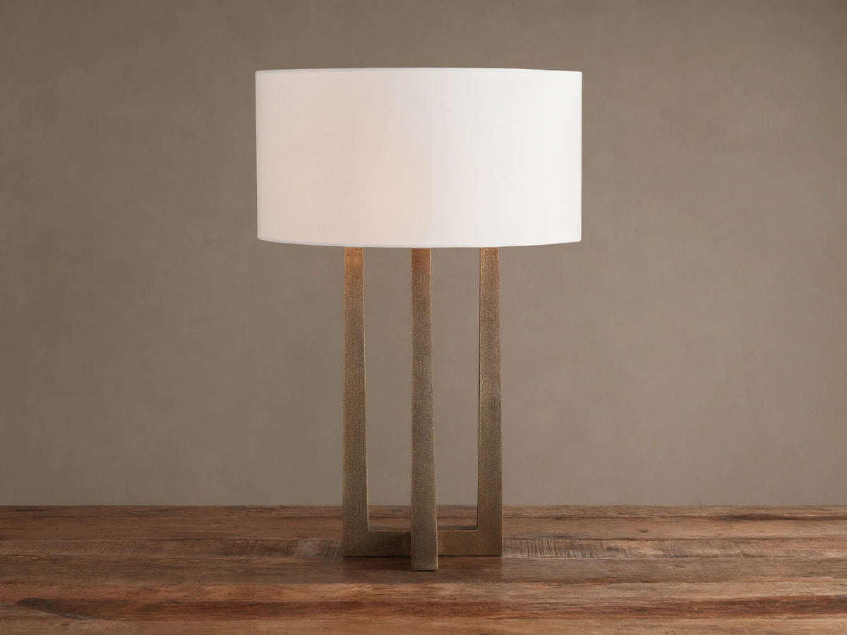 Emmet Table Lamp: lit white cylindrical linen shade, textured antiqued brass three-legged base, on a rustic wood table.