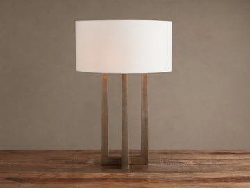 Emmet Table Lamp: lit white cylindrical linen shade, textured antiqued brass three-legged base, on a rustic wood table.