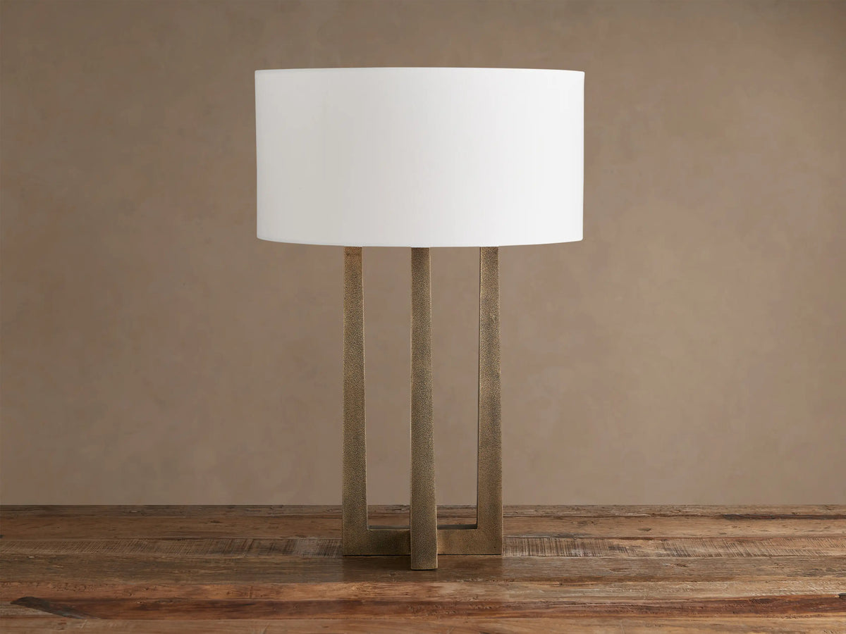 Emmet Table Lamp: lit white cylindrical linen shade, textured antiqued brass three-legged base, on a rustic wood table.
