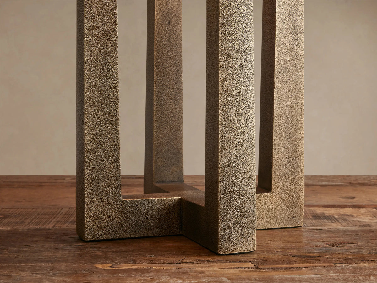 Emmet Table Lamp with a white cylindrical shade & three antiqued brass rectangular supports on a rustic wooden table.