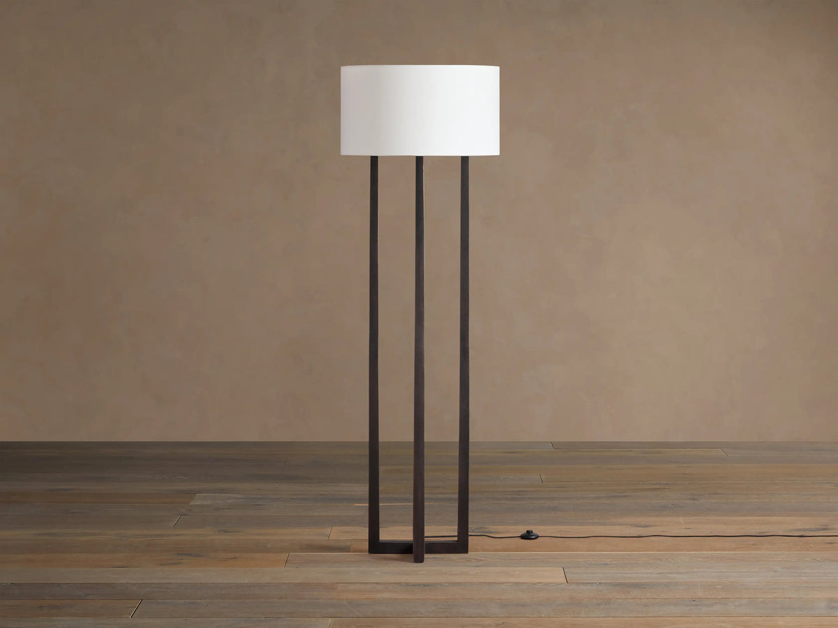 Emmet Floor Lamp with dark antiqued bronze/brass finish, three vertical rectangular bars, white cylindrical linen shade, on wood floor. Foot switch.