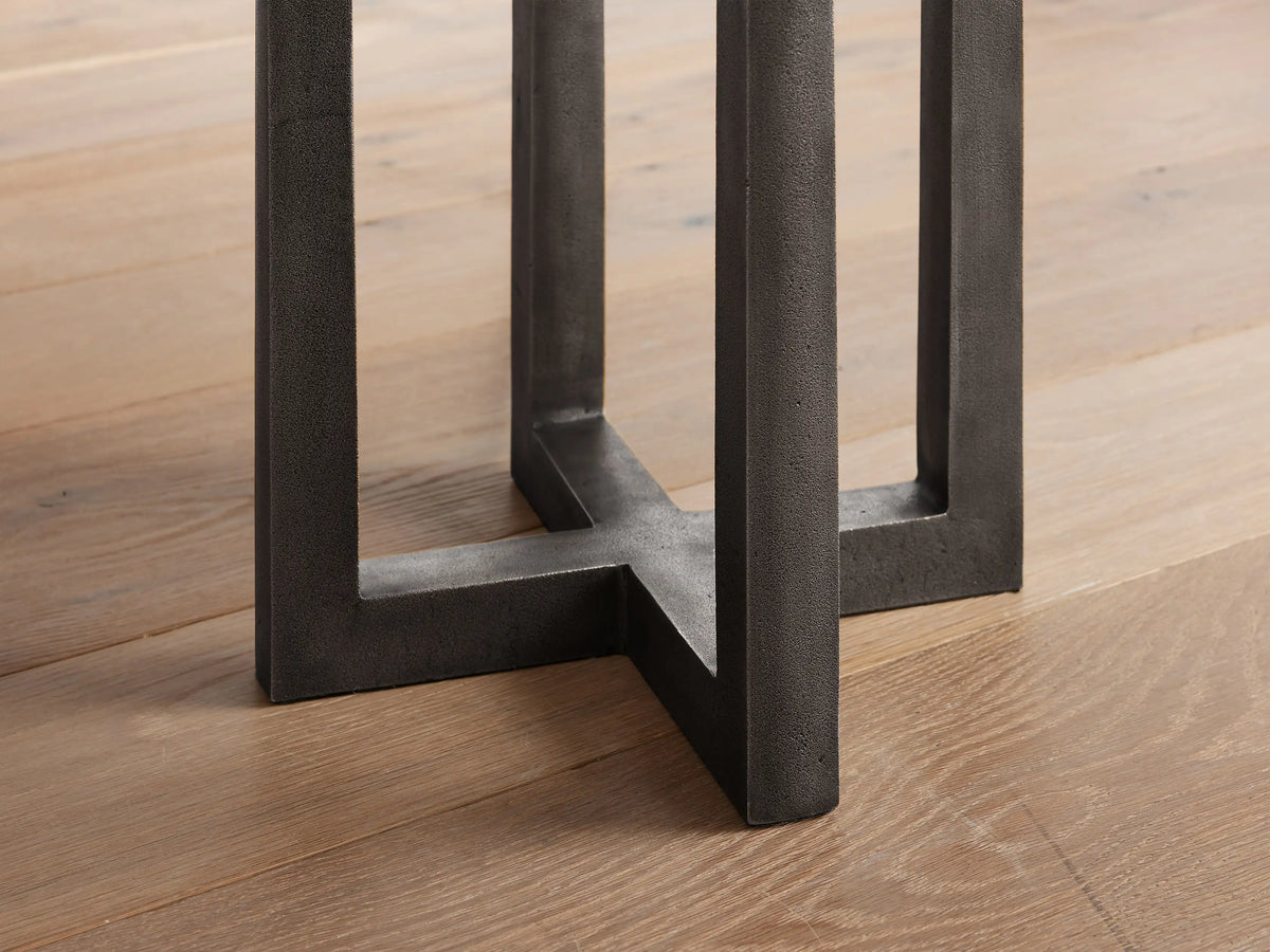 Close-up of Emmet Floor Lamp's dark, textured, antiqued brass base with geometric cross pattern resting on a light wooden floor.