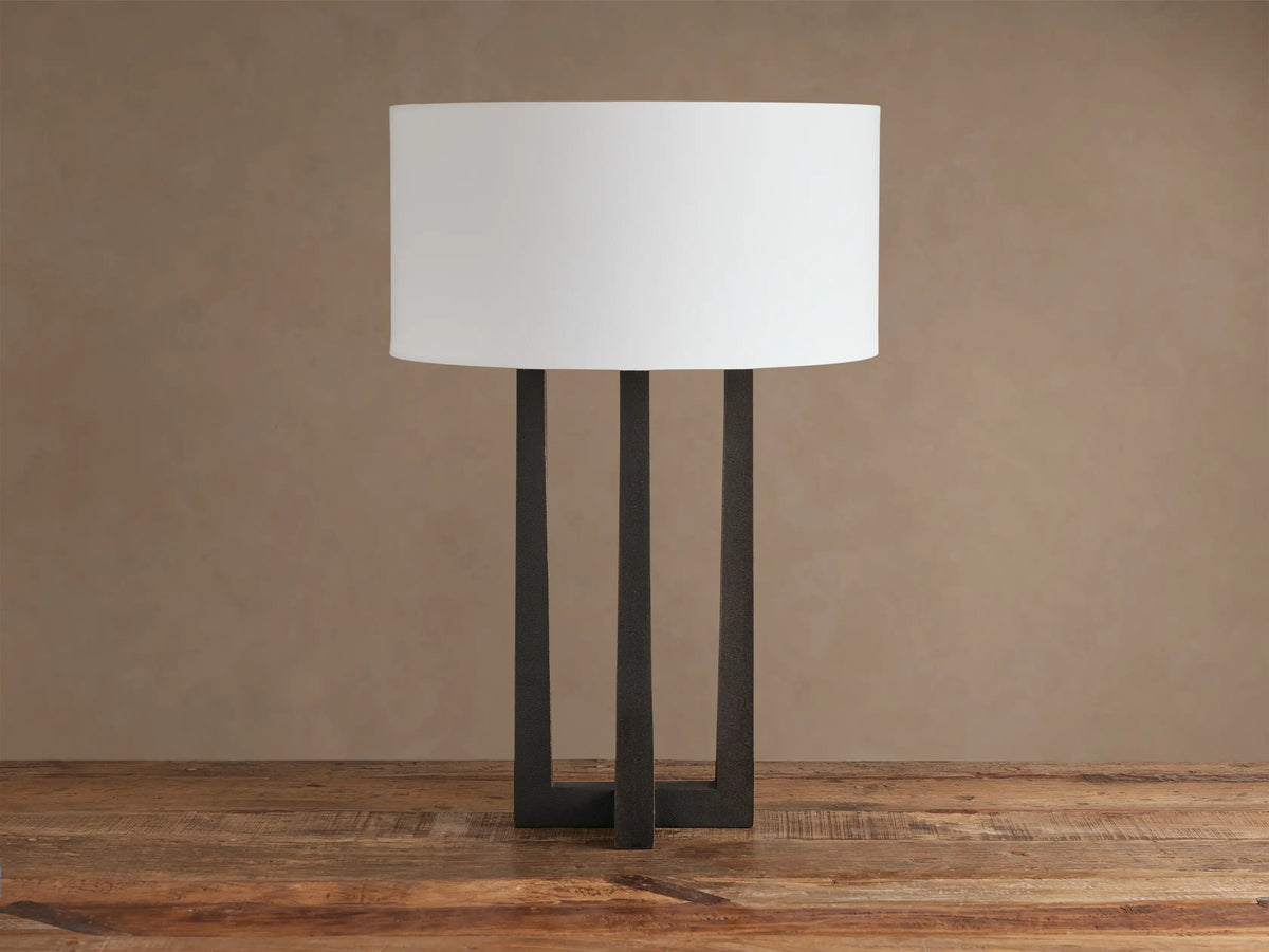 Emmet Table Lamp with white linen shade and dark, textured bronze three-leg base, sitting on a rustic wood table.