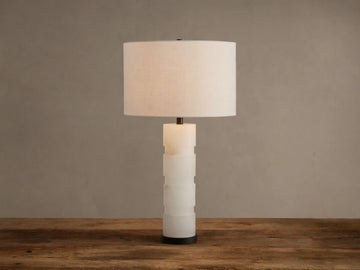 Novalie Table Lamp: Carved white alabaster body with bronze base, topped by a large off-white fabric drum shade on a wooden surface.