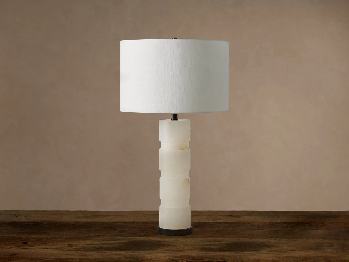 Novalie Table Lamp: Carved white alabaster body with bronze base, topped by a large off-white fabric drum shade on a wooden surface.